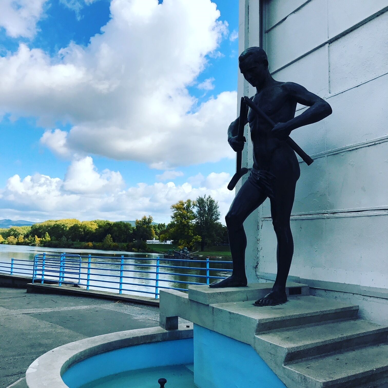 Una estatua de una persona sosteniendo un murciélago, cerca de una masa de agua con una baranda azul y escalones que bajan hasta el agua.