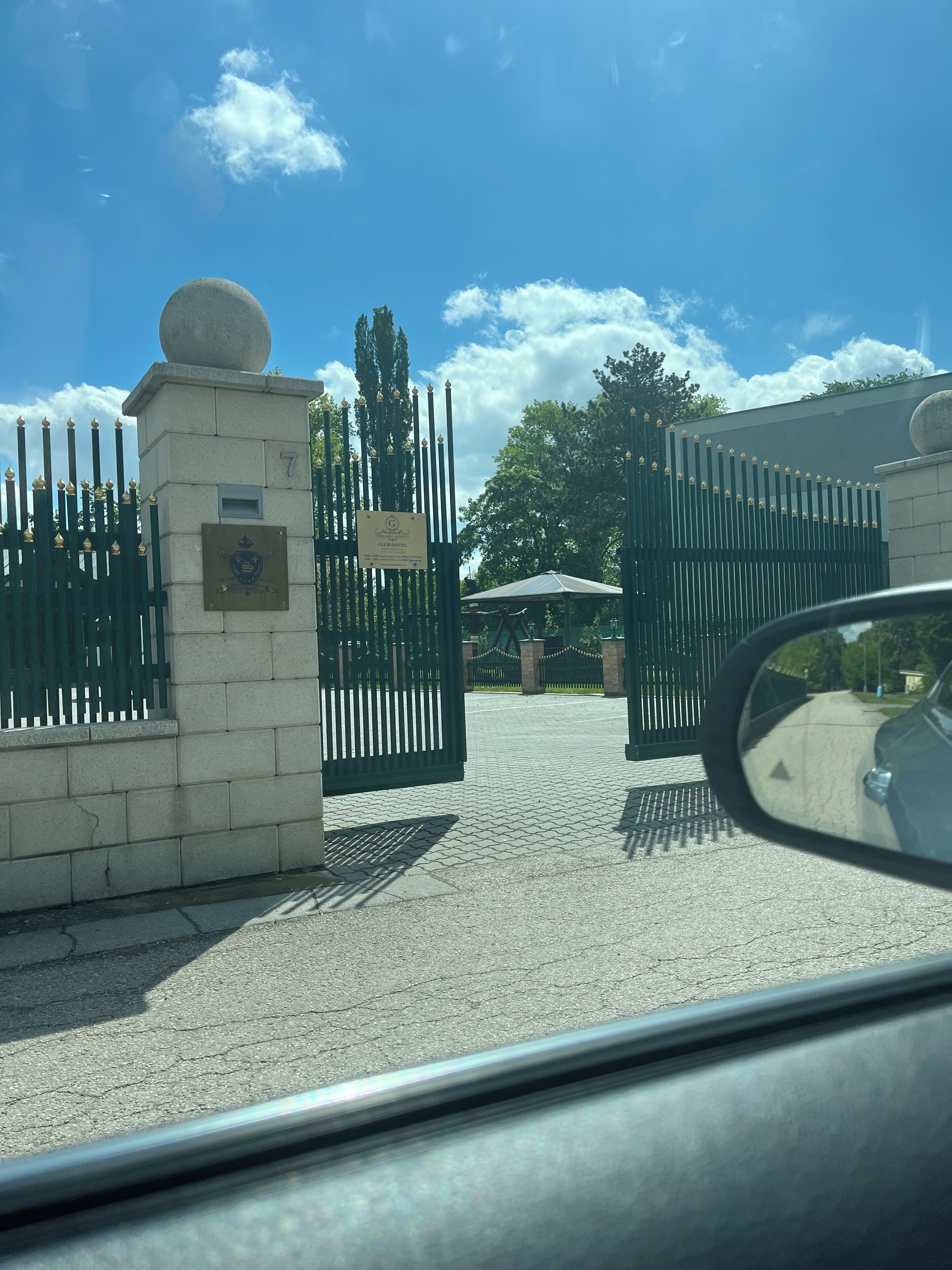 A gated entrance with a plaque, a clear sky, and a car's side mirror reflection.