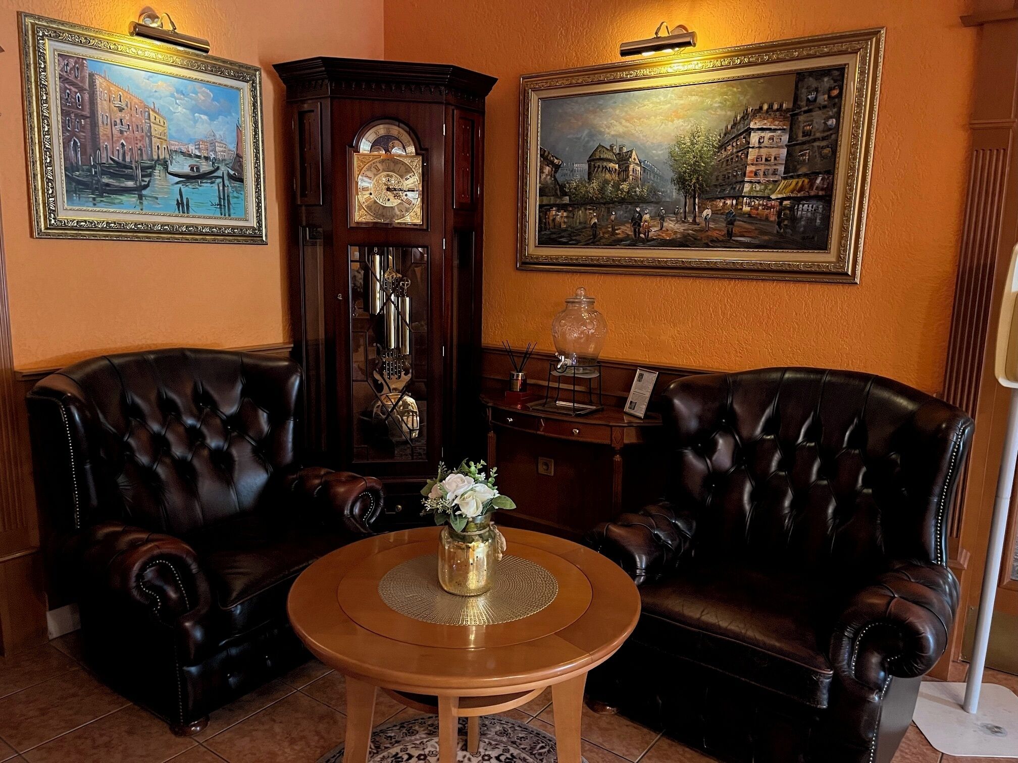 Una habitación con dos sillones de cuero, una mesa de centro de madera con un florero, un reloj de pie y dos cuadros enmarcados en las paredes.