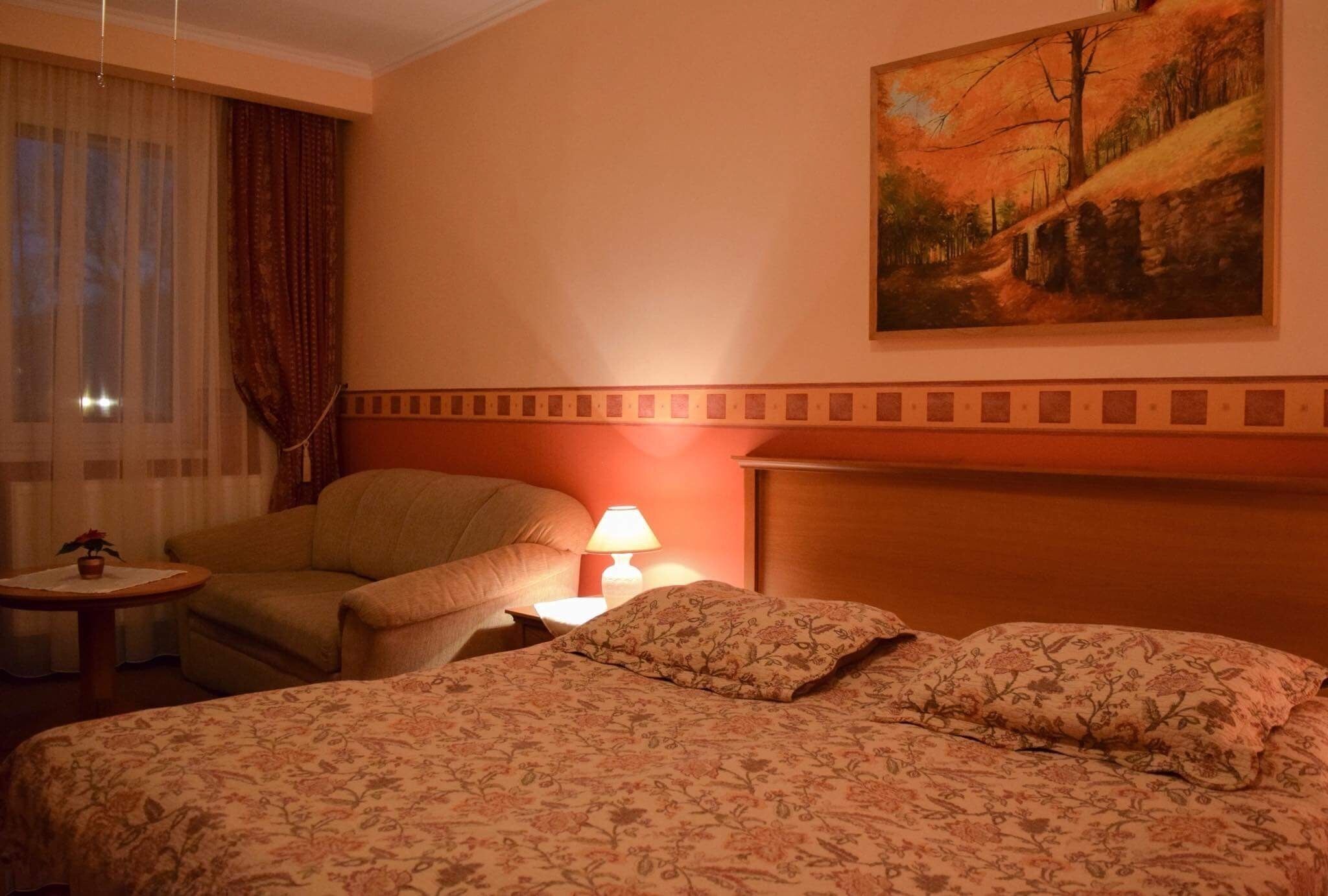 A hotel room with a bed, a chair, a small table with a lamp, and a painting on the wall.