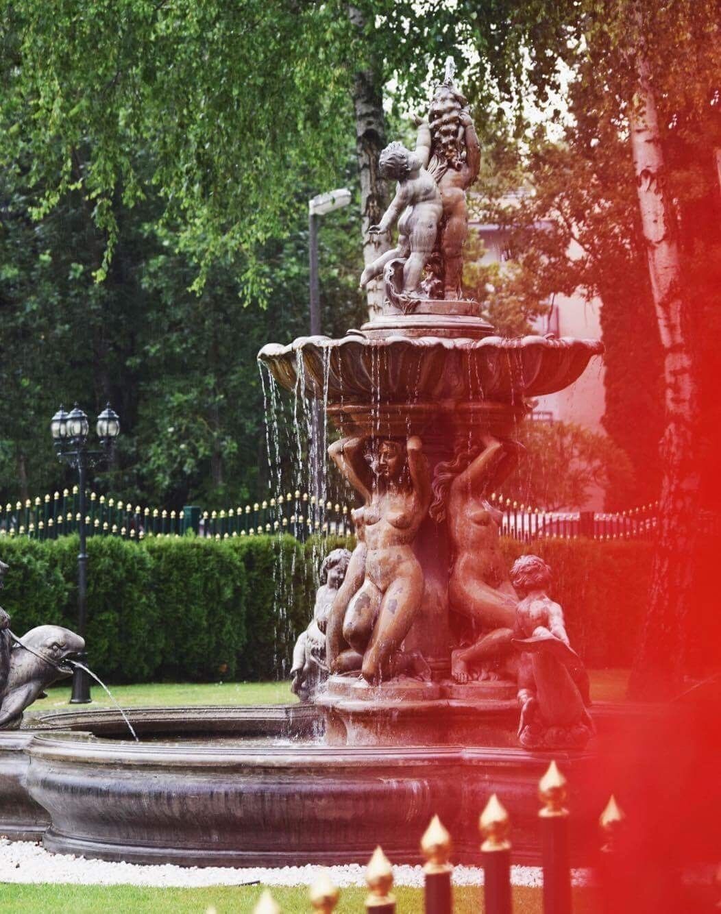 A tiered fountain with multiple statues, surrounded by a hedge and street lamps.