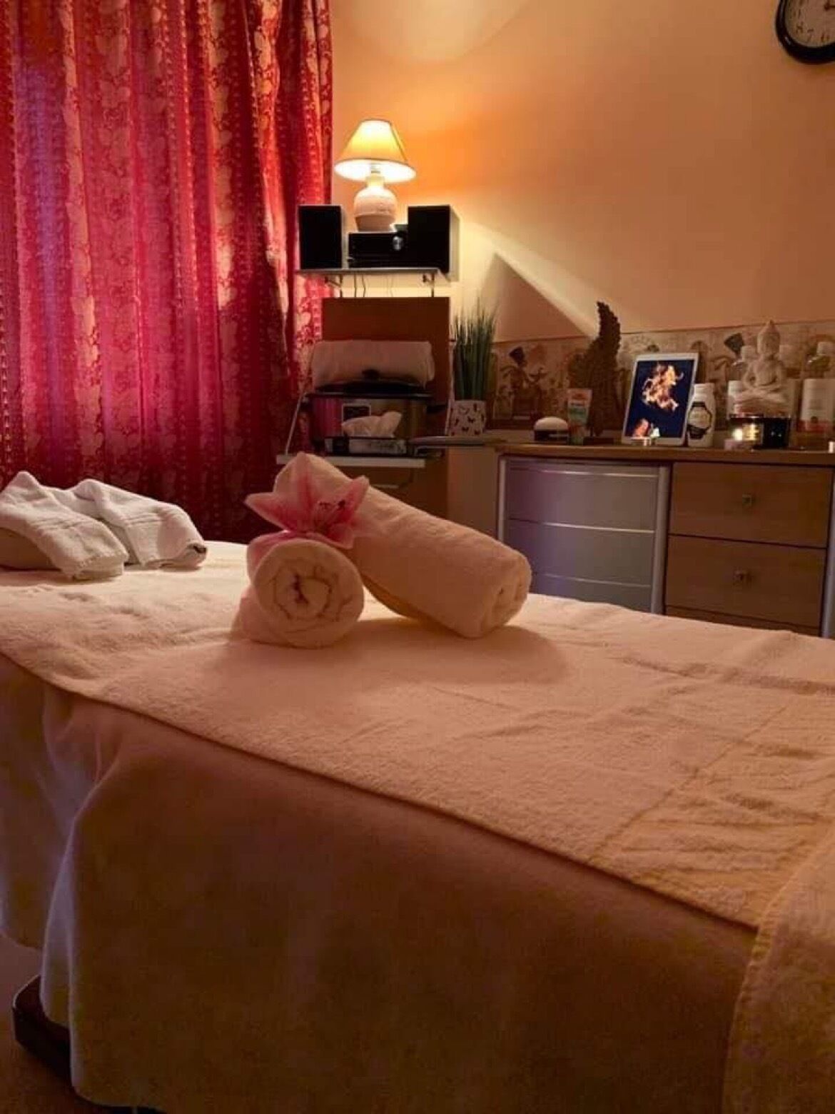 Ein ordentlich bezogenes Bett mit Handtüchern und einem Zierkissen, eine Nachttischlampe und eine Uhr an der Wand.