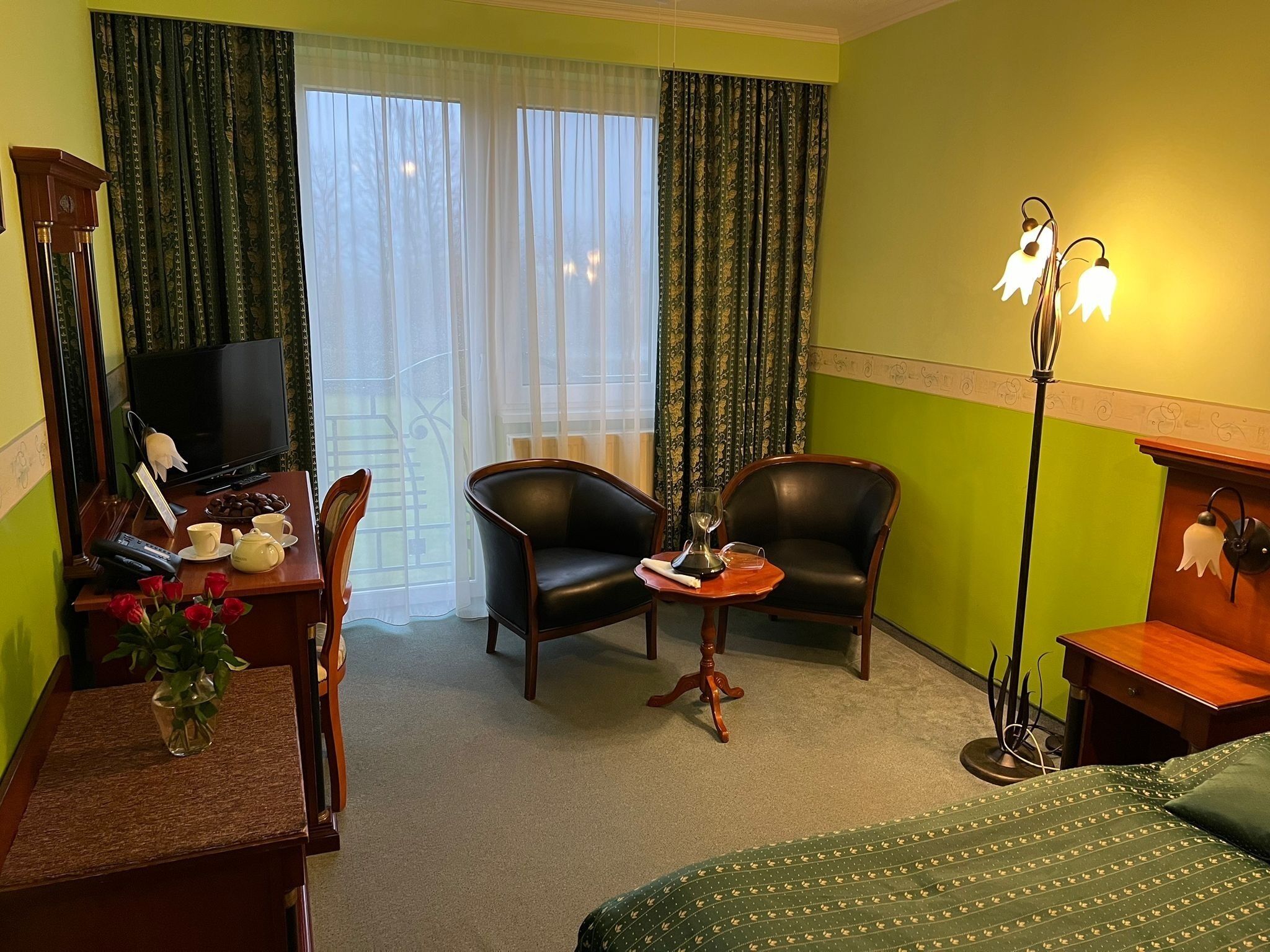 A hotel room with a bed, two chairs, a desk with a telephone and a vase of flowers, a television, and a lamp.