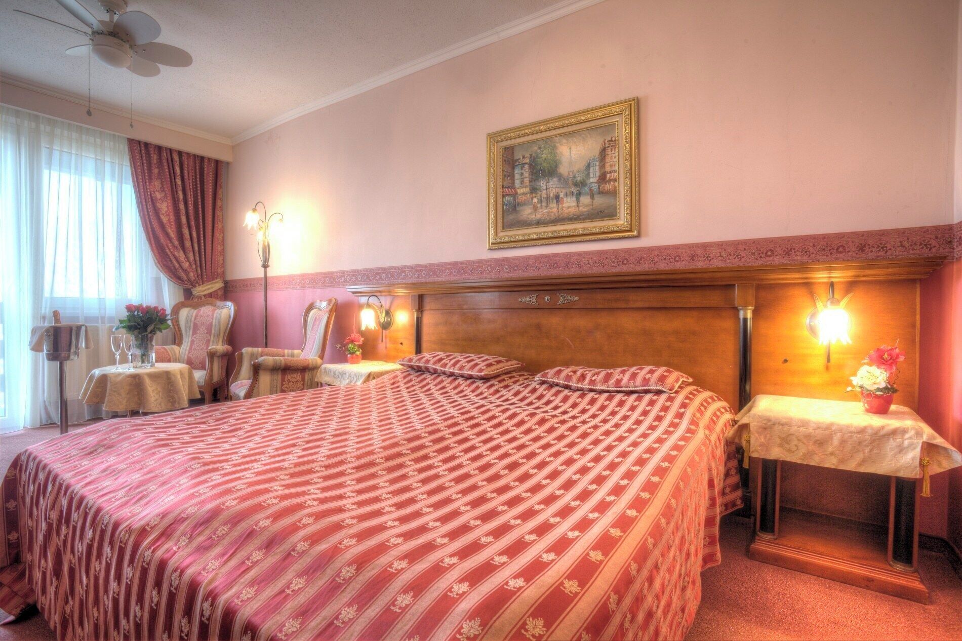 A hotel room with a large bed, two armchairs, a small table with a vase of flowers, and a painting on the wall.