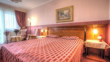 A hotel room with a large bed, two armchairs, a small table with a vase of flowers, and a painting on the wall.