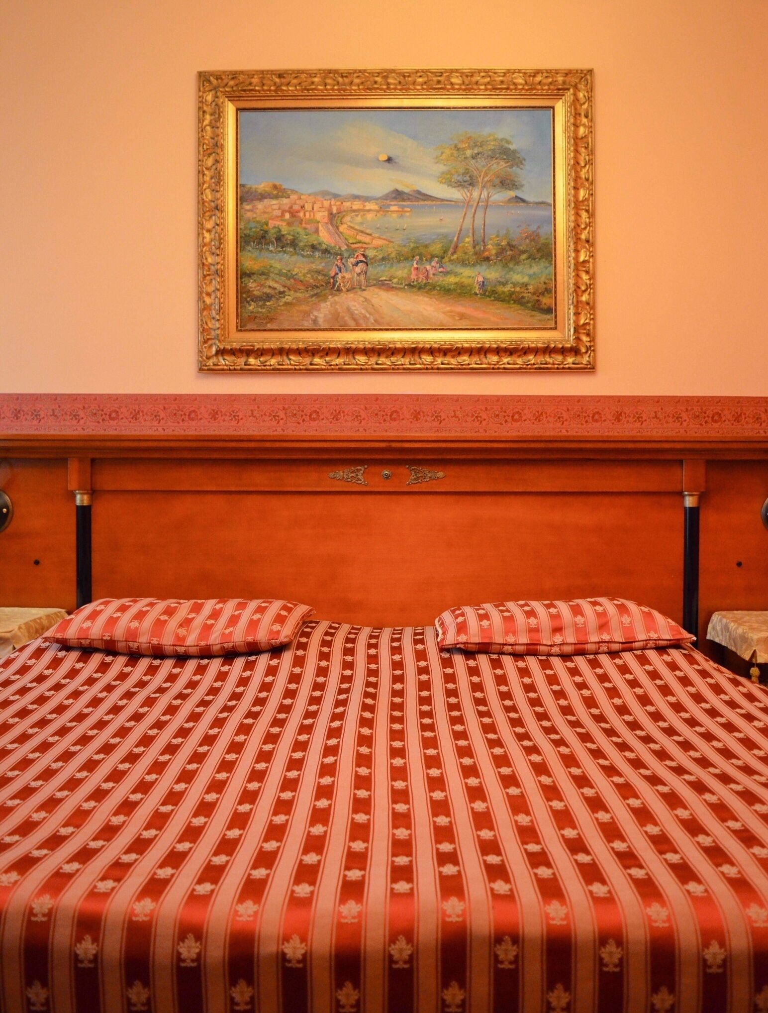 Een hotelkamer met een bed, voorzien van een rood-witte strepen dekbedovertrek, een houten hoofdbord en een lijst met een landschapsdecoratie erboven.