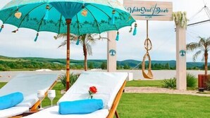 Twee ligbedden met blauwe kussens en een turquoise parasol, een bord met de tekst 'Wake/Soul Beach' en palmbomen op de achtergrond.