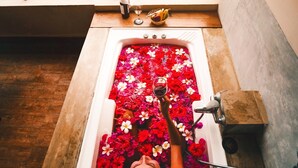 Eine Person entspannt sich in einer Badewanne voller roter Blüten, mit Blick auf einen Pool und Palmen.