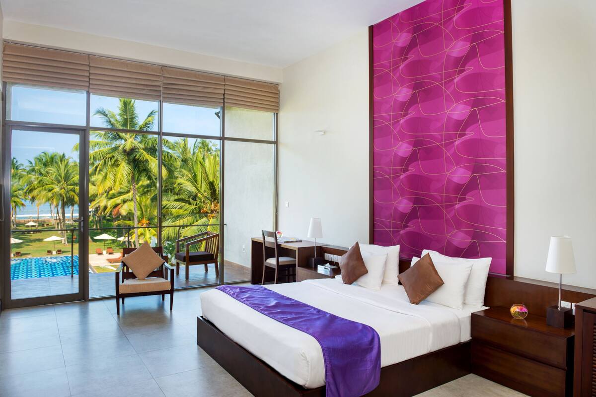 A hotel room with a large window, a bed with a purple throw, a desk, and a chair.