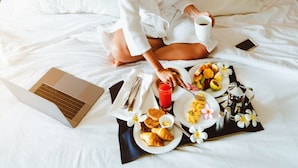 Eine Person sitzt auf einem Bett, vor sich ein Laptop und ein Tablett mit Frühstücksgegenständen. Im Hintergrund hängt ein Gemälde.