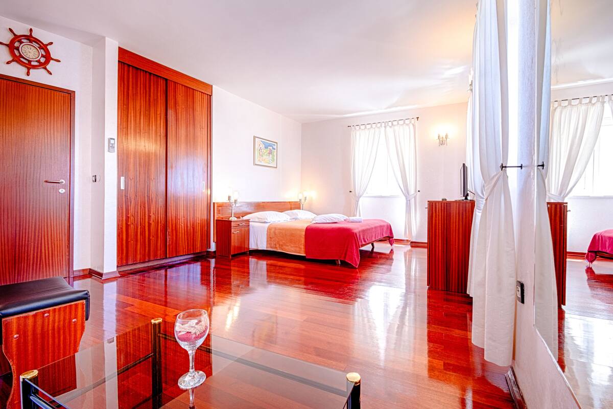 A hotel room with a wooden floor, a bed with white and red bedding, a wooden wardrobe, a glass table with a wine glass, and a ship's wheel on the wall.