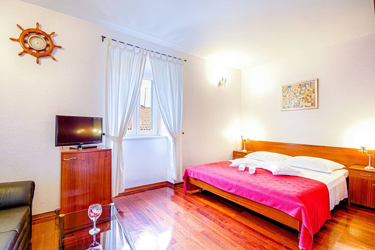 A hotel room with a wooden bed, a television on a cabinet, a window with white curtains, and a wall clock.