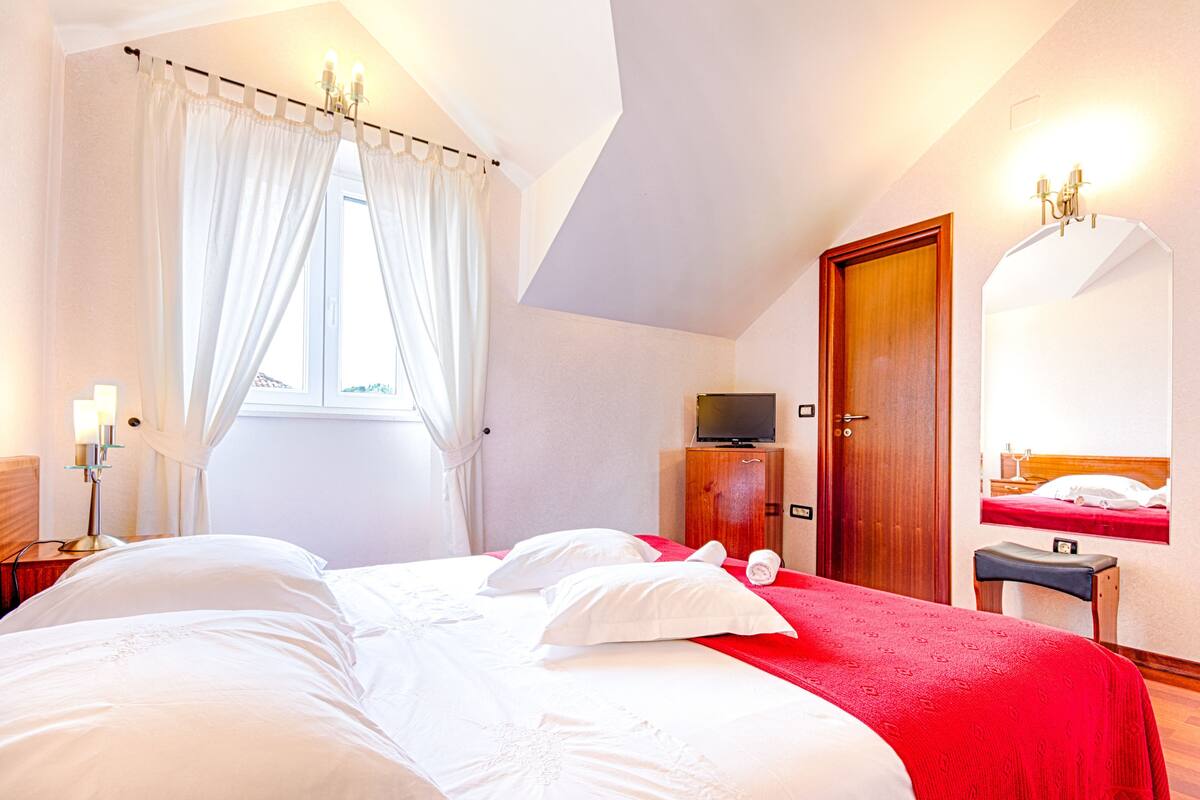 A hotel room with a large bed, a television on a wooden cabinet, a mirror on the wall, and a window with curtains.