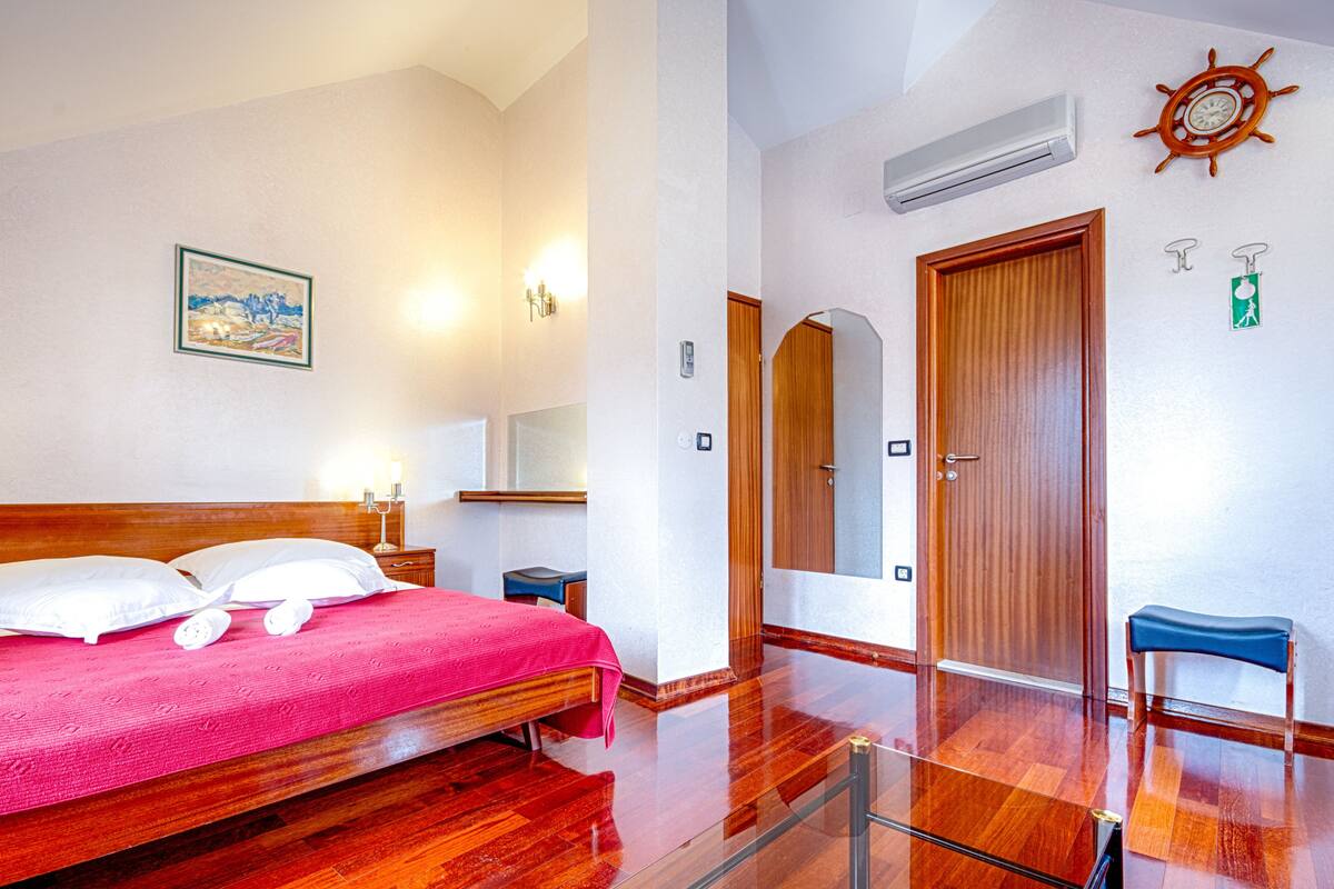 A hotel room with a wooden bed, a bedside table, a nightstand, a mirror, a wall-mounted air conditioner, a door, and a bench.