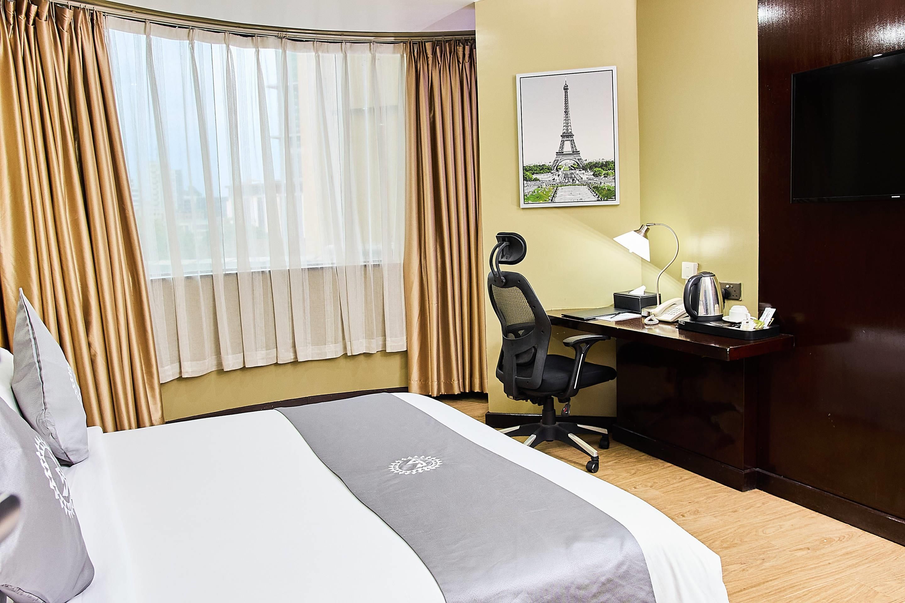 Ein Hotelzimmer mit einem Bett, einem Schreibtisch mit Stuhl, einer Lampe, einem Telefon und einem Bild des Eiffelturms an der Wand.