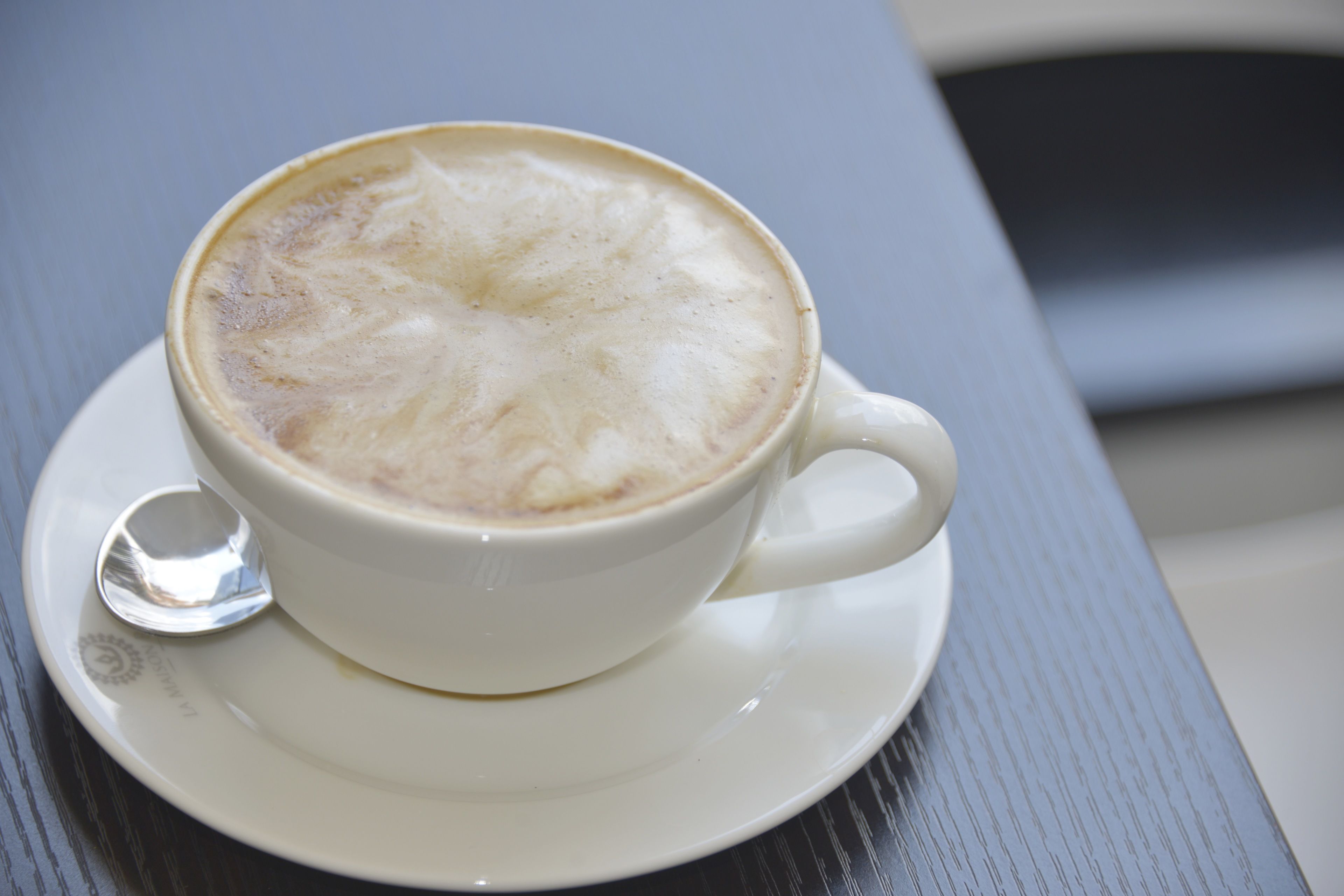 Une tasse de café surmontée d’une mousse onctueuse, posée sur une soucoupe blanche, elle-même posée sur une table.