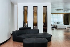 A modern living room with a sectional sofa and ottomans, a large window with curtains, and a visible kitchen area. - La Maison Royale - Westlands (Nairobi)