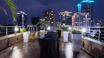 Una terraza en la azotea con mobiliario exterior, plantas en macetas y el horizonte de la ciudad de noche.