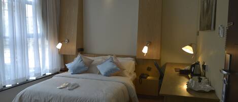 Habitación de hotel con cama, lámparas de noche, escritorio con teléfono y tetera, y un cuadro en la pared.