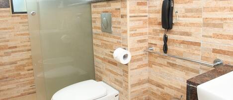 Un baño con inodoro, lavamanos, cabina de ducha de vidrio, toallero y teléfono.