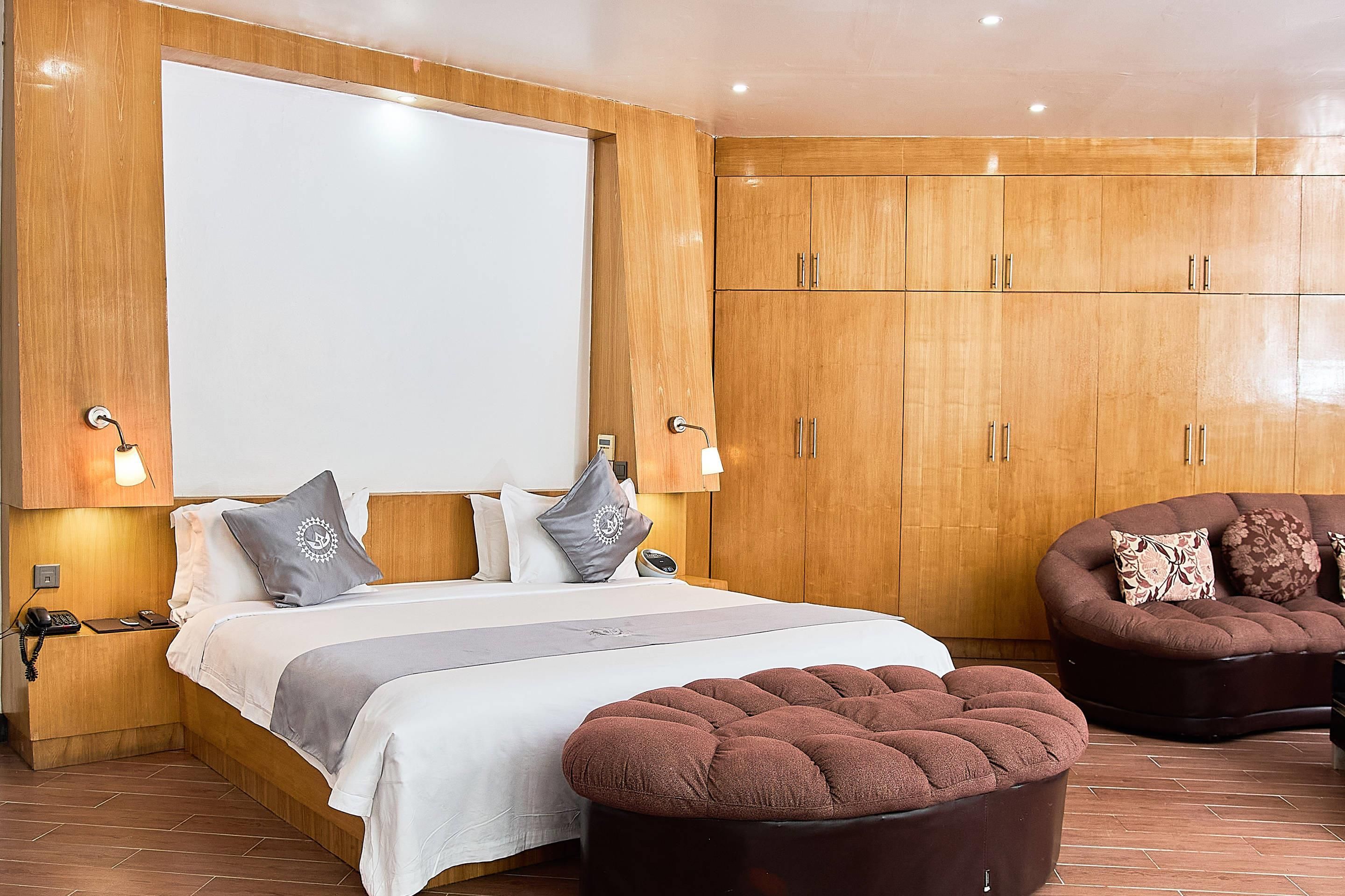 A hotel room with a large bed, wooden wardrobe, two bedside lamps, a brown ottoman, and a brown sofa.