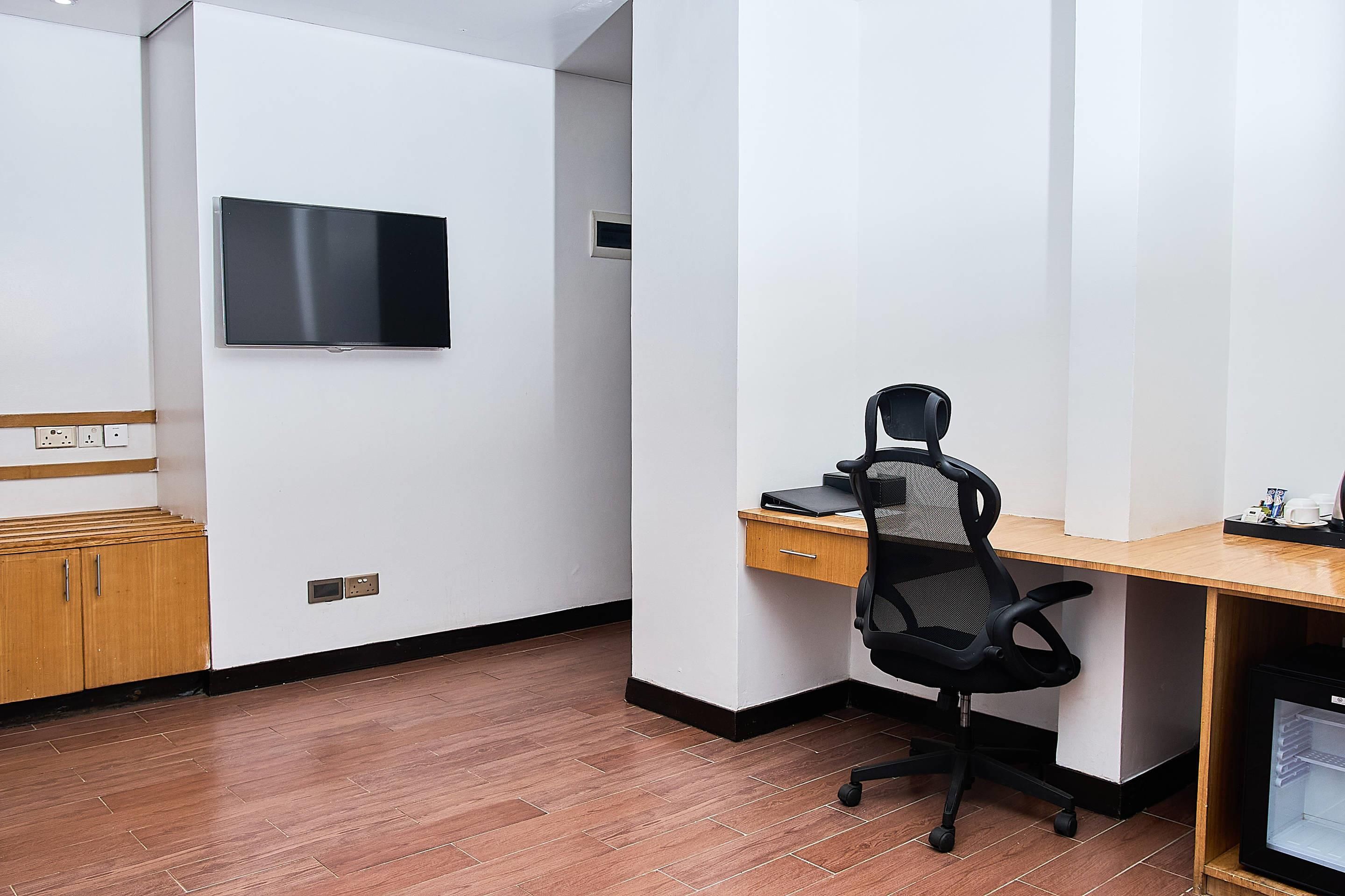 A room with a wooden desk, a black office chair, a television mounted on the wall, and a small cabinet.