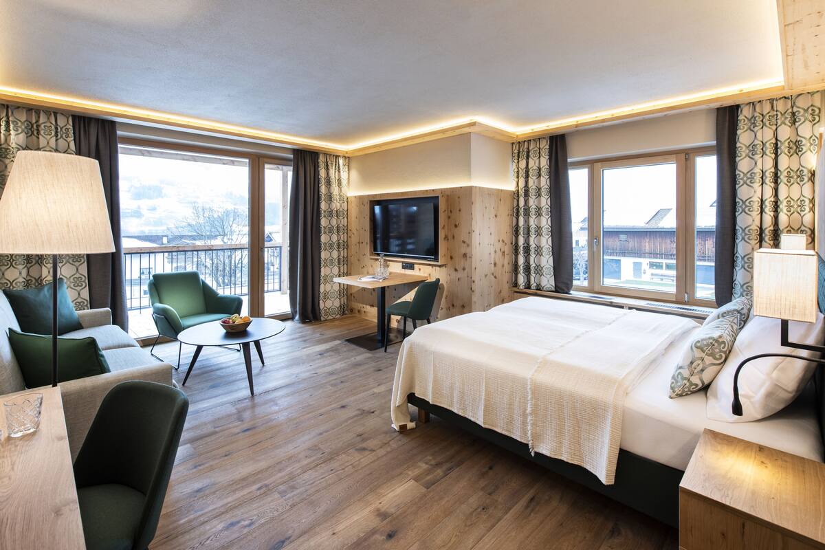 A hotel room with a bed, a sofa, a desk, a TV, and large windows.