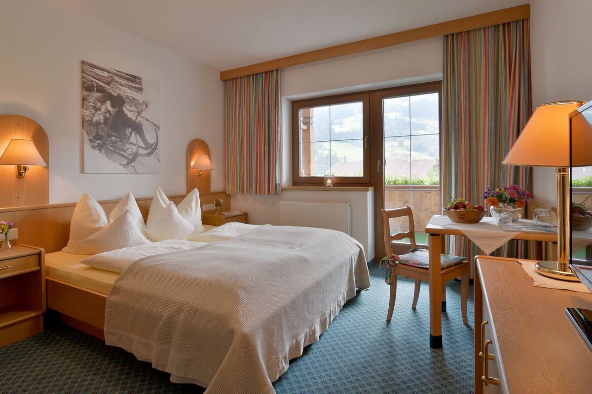 A hotel room with a bed, bedside lamps, a desk with a chair, a window with curtains, and a view of the outdoors.