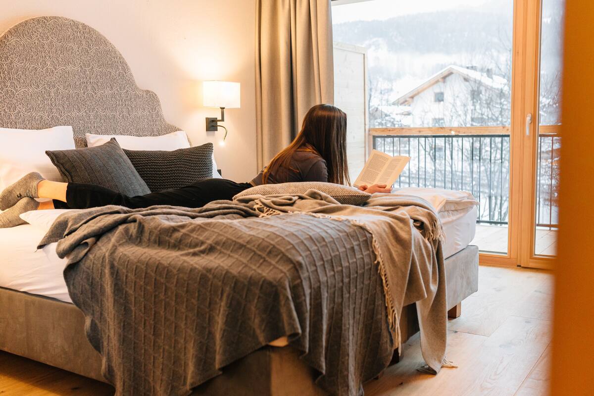 A bedroom with a bed, pillows, a blanket, and a person reading a book.