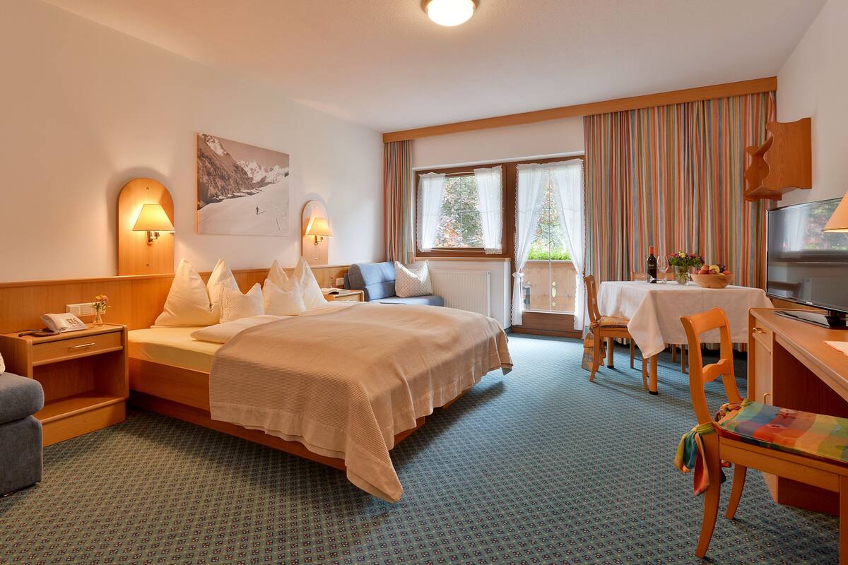 A hotel room with a bed, a desk with a TV, a chair, a small table, a window with curtains, and a painting on the wall.