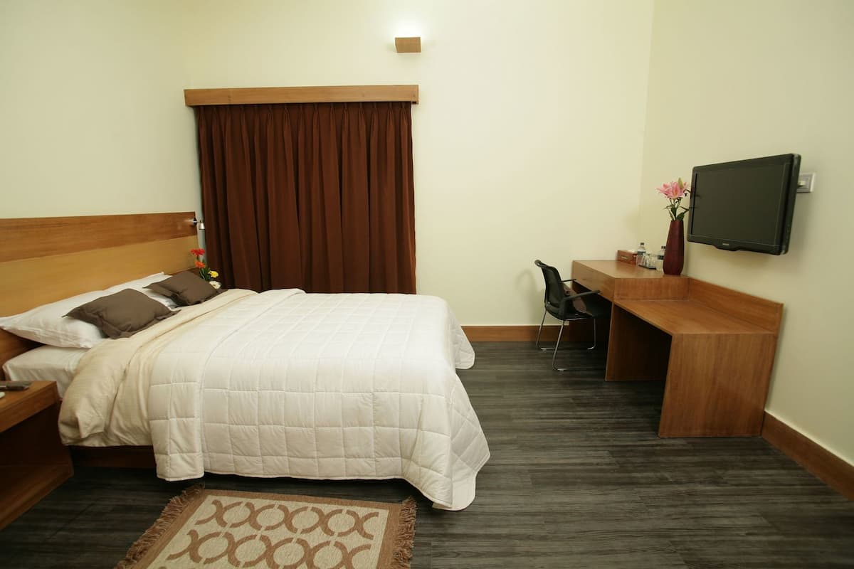 Habitación de hotel con cama, escritorio, silla, televisión y una pequeña mesa con un jarrón.