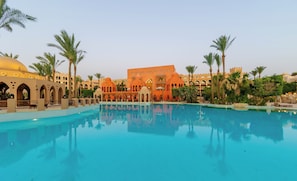 A large pool with clear blue water, surrounded by palm trees and a building with arches and domes.