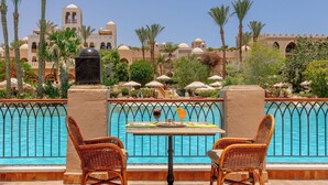 Un espace aménagé au bord de la piscine avec des fauteuils en osier et une table, une piscine dotée d’une rambarde en métal, et un bâtiment à l’architecture caractérisée par des dômes en arrière-plan.