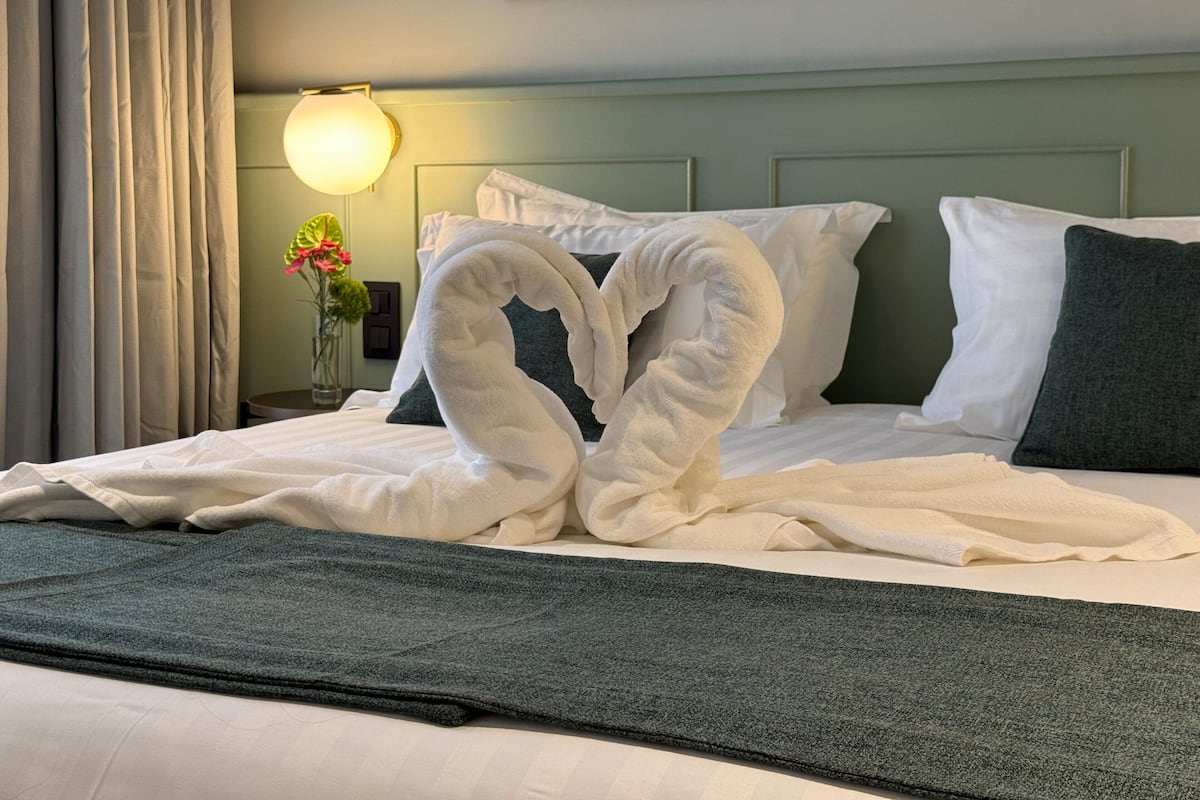 Een net opgemaakt bed met witte en groene kussens, een groene plaid en een vaas met bloemen op het nachtkastje.