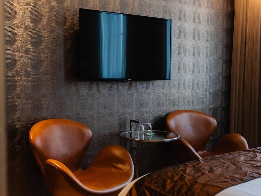Une chambre d’hôtel équipée d’un téléviseur à écran plat fixé au mur, de deux fauteuils en cuir, d’une petite table avec des verres et d’un lit recouvert d’une courtepointe à motifs.