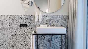 A modern bathroom with a granite backsplash, a round mirror, a sink with a towel rack, and a shower area.