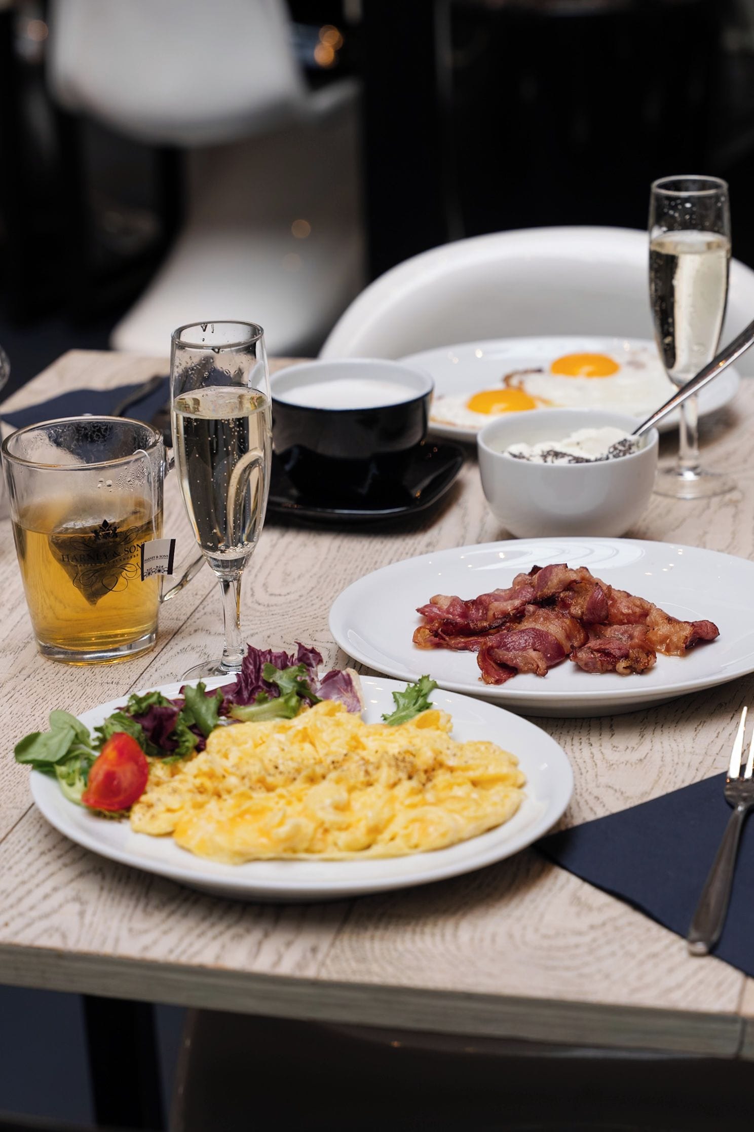 A breakfast table with plates of eggs, bacon, and a salad, two glasses of water, and a mug of tea.