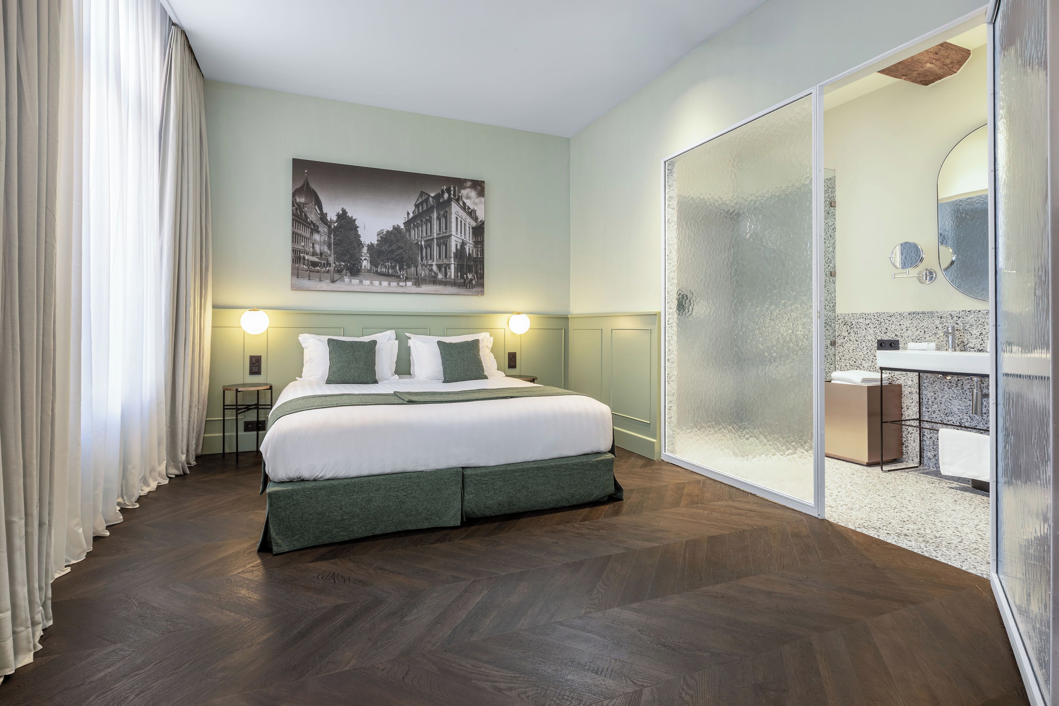 Una habitación de hotel con una cama, una mesita de noche, una lámpara de pared, un cuadro y una puerta corrediza de vidrio que da a un baño.