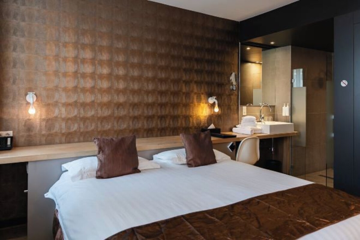 Ett hotellrum med en stor säng, en sänggavel i trä, en spegel, ett skrivbord med en stol och ett badrum med handfat och spegel.