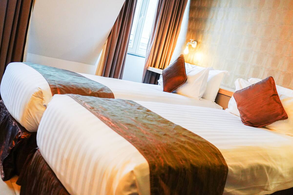 Een hotelkamer met twee bedden, elk voorzien van witte lakens en bruine hoofdborden. Daartussen staat een nachtkastje met een lamp.