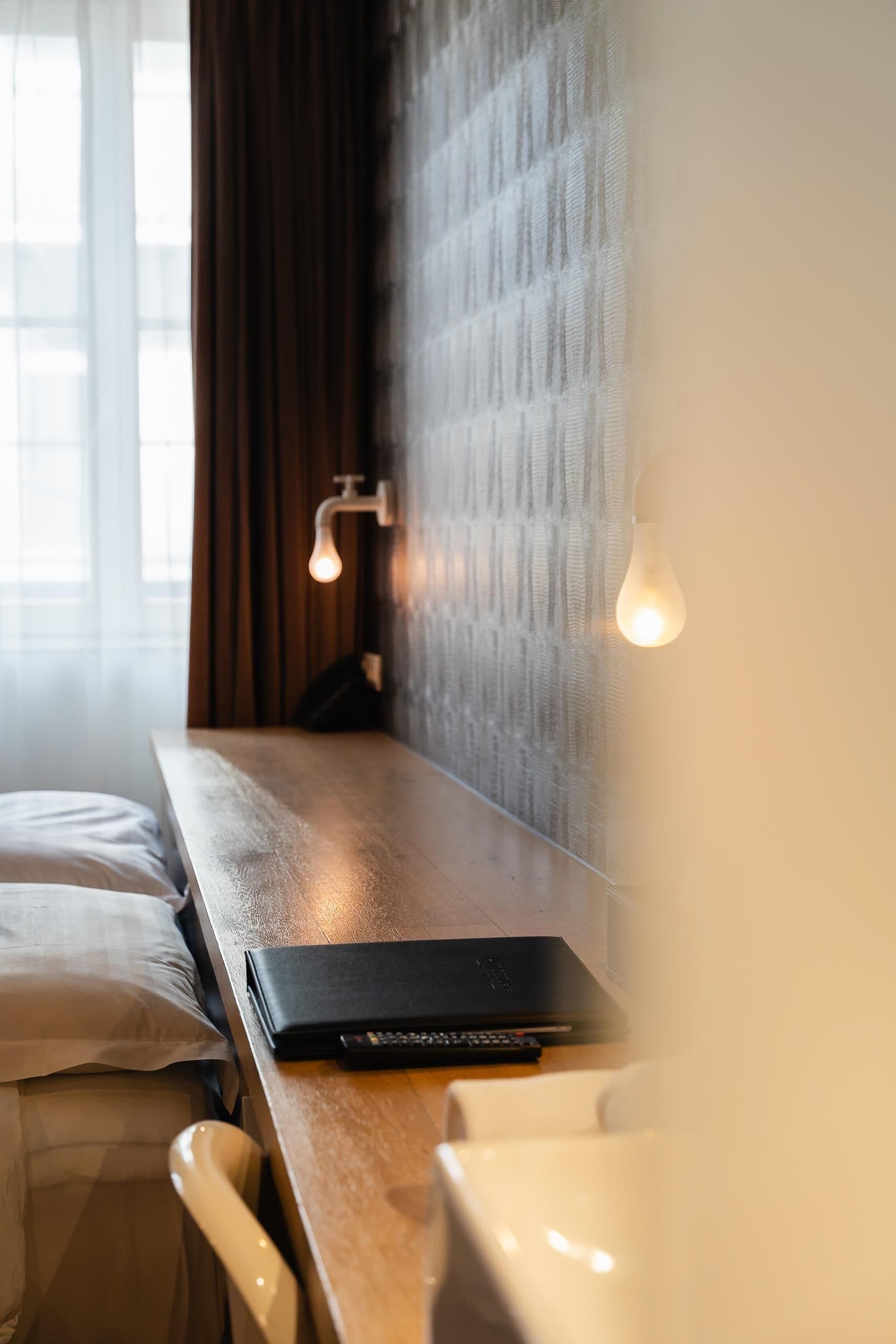 A hotel room with a wooden desk, a laptop, a remote control, and a bed with a pillow.