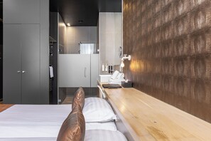 Habitación de hotel moderna con una cama, una mesita de noche de madera, un espejo y un baño con ducha y toallero.