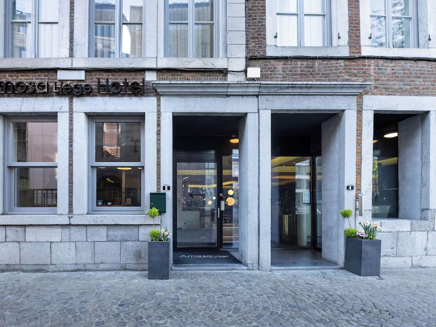 Entrada de hotel con puerta de vidrio, puertas numeradas y una pequeña maceta exterior.