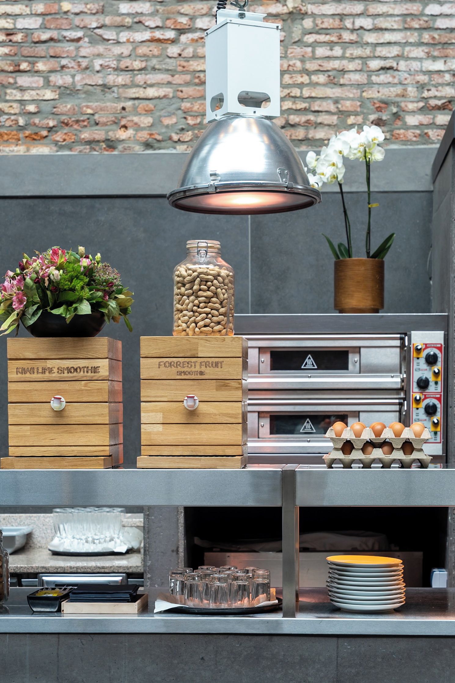 Una zona de cocina con fondo de pared de ladrillo, una lámpara colgante, una caja de madera con la etiqueta 'FORREST FRUIT SMOOTHIES', un tarro de cacahuates, una orquídea en maceta y una pila de platos.