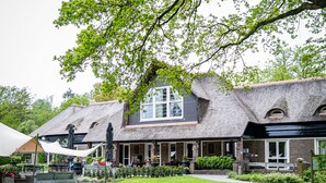 Een traditioneel gebouw met rieten dak, een centraal raam en een goed onderhouden tuin met gazon en bloembedden.