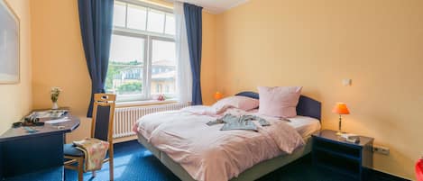 A hotel room with a bed, a desk, a chair, a window with curtains, a nightstand with a lamp, and a radiator.