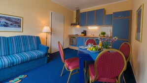 Een knus woongedeelte met een bank, eettafel, stoelen, kitchenette en een schilderij aan de muur.