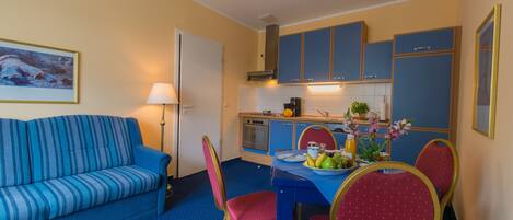 A compact living space with a sofa, dining table, chairs, kitchenette, and a painting on the wall.