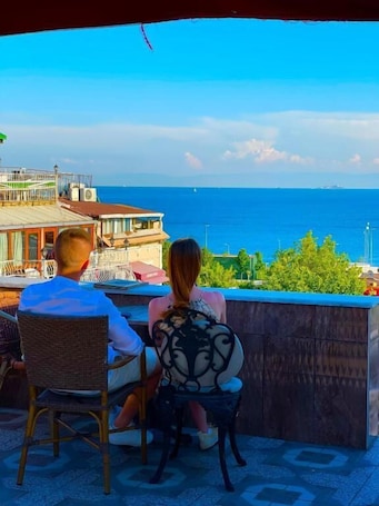 A couple seated at an outdoor table with a view of the sea and buildings in the background.