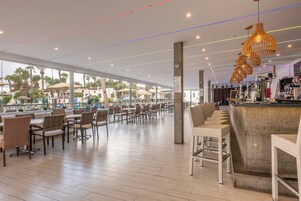 A spacious indoor area with tables and chairs, a bar area with a counter, and a view of palm trees outside.